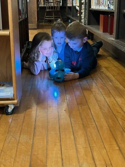 Three children play with a glowing blue toy on a wooden library floor surrounded by bookshelves